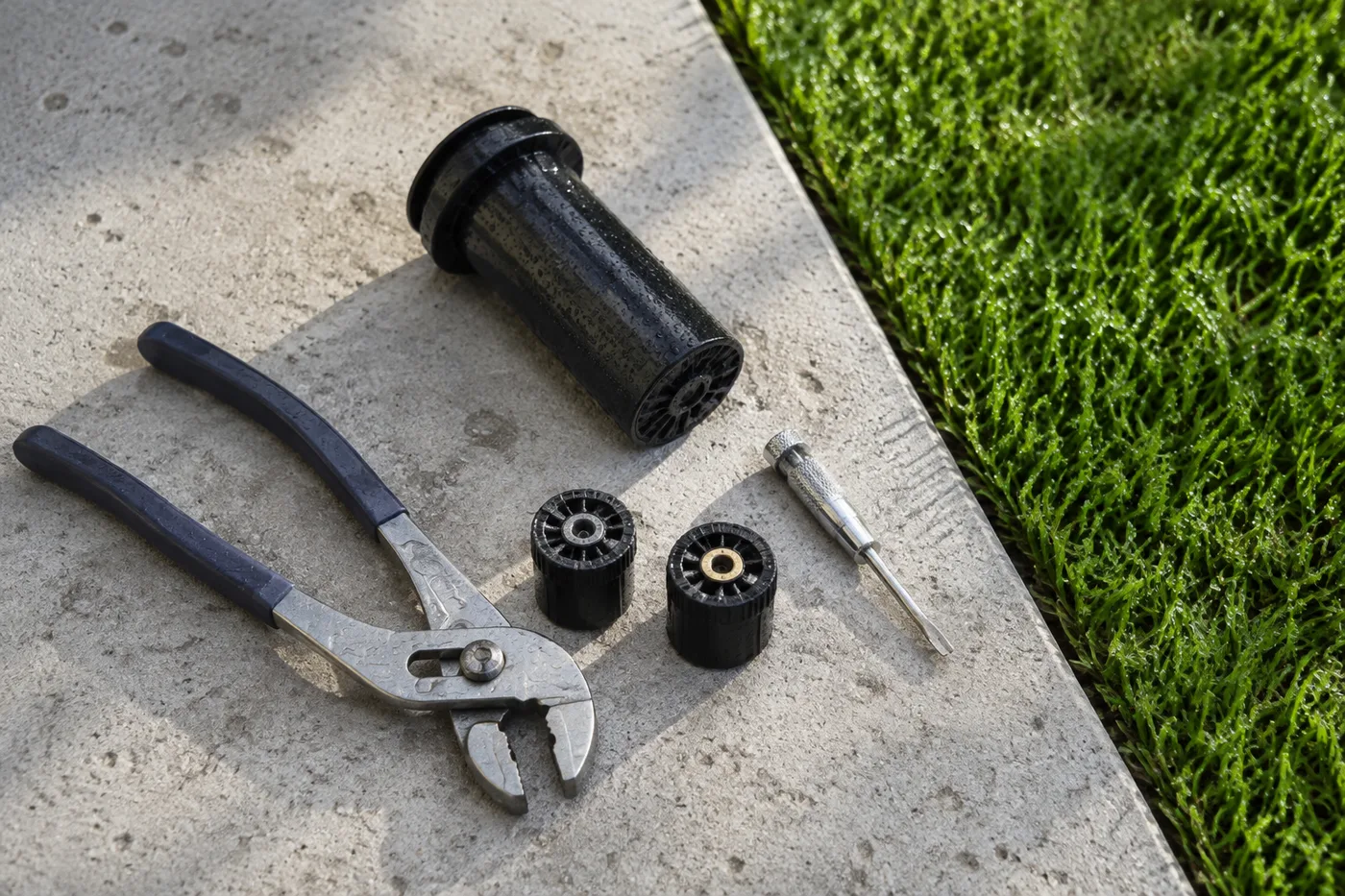 Sprinkler head, spray nozzles, pliers and a small screwdriver laid out on wet concrete beside green turf